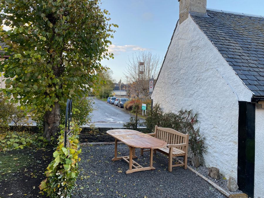 Teak table and bench on the gravel patio beside the cottage's white gable, autumn leaves on the ground
